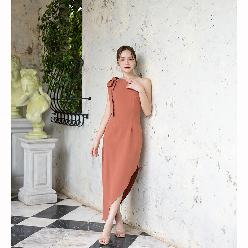 kudapy เดรสยาสีสีน้ำตาลอมส้ม/สีครีม ช่วงบนเป็นแบบปาดไหล่ข้างเดียวแต่งผ้าพับอกดูไม่เรียบจนเกินไป มีสายผูกโบว์ที่ไหล่ด้านขวาเพิ่มความหวาน